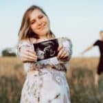 pregnant woman holding ultrasound photo in field with husband behind