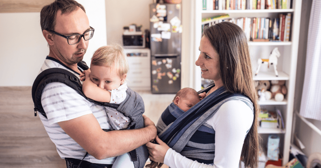 Two parents wearing their babies in a carrier and a wrap sling