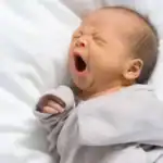 Newborn baby yawning while lying on a white blanket.