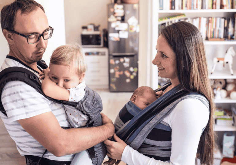 Two parents wearing their babies in a carrier and a wrap sling