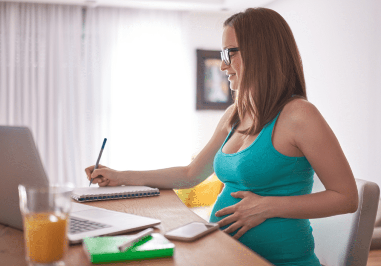 Pregnant woman at desk with laptop writing on checklist
