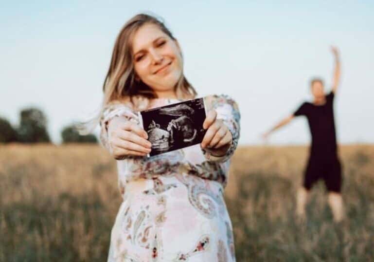 pregnant woman holding ultrasound photo in field with husband behind