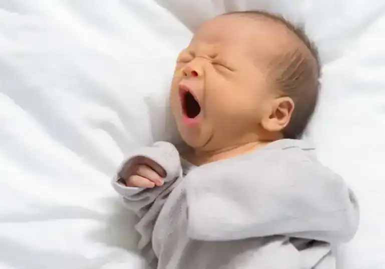 Newborn baby yawning while lying on a white blanket.